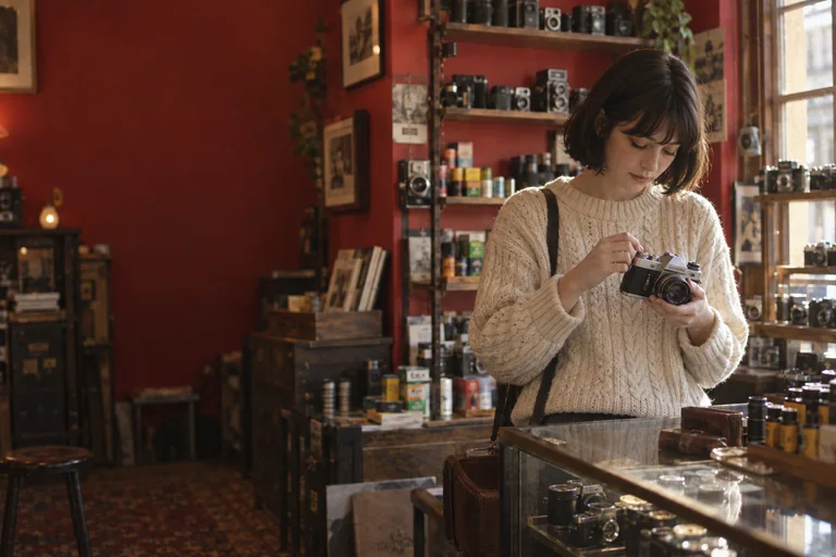 A woman in a cream knit jumper winding on a vintage film camera in a red-walled camera shop
