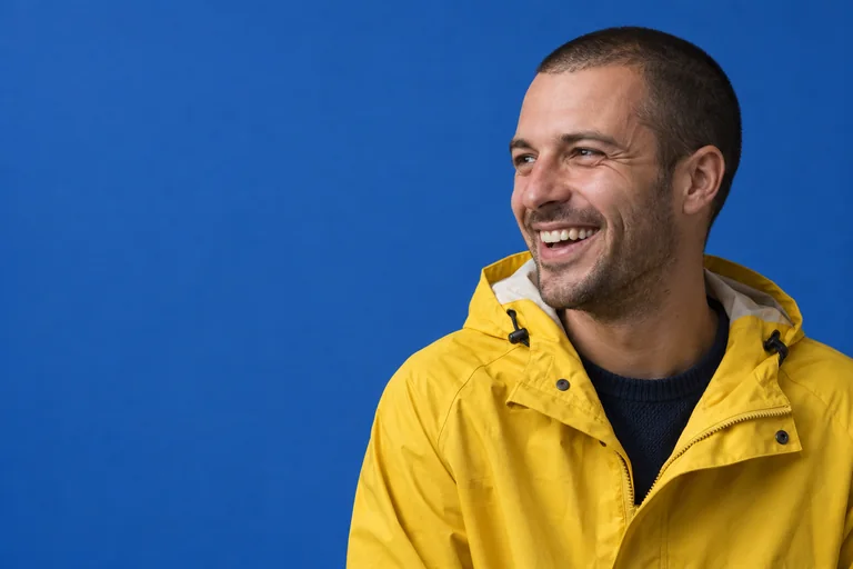 A man in a yellow rain jacket laughing against a cobalt blue backdrop
