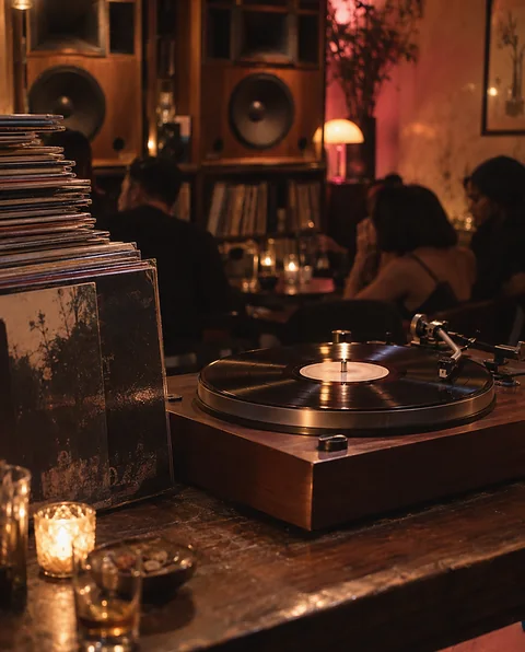 A turntable playing a record on the counter of a dim listening bar, with vintage speakers and listeners behind